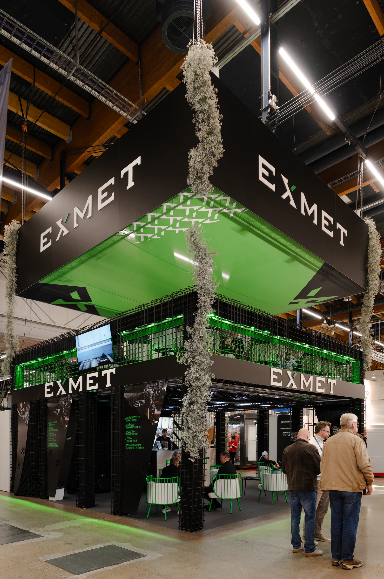 Two-level Exmet Group stand at FinnBuild with green-lit ceiling and hanging greenery